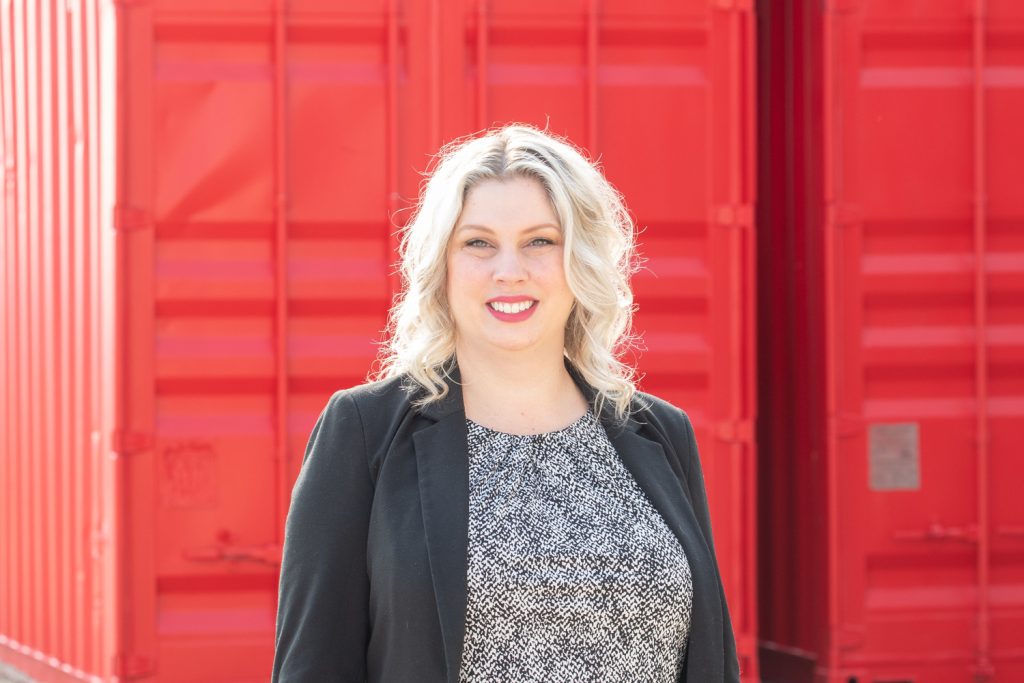 Une femme à la peau claire et aux cheveux blonds ondulés, vêtue d’un blazer noir et d’un chemisier noir et blanc, sourit devant des conteneurs maritimes rouges.